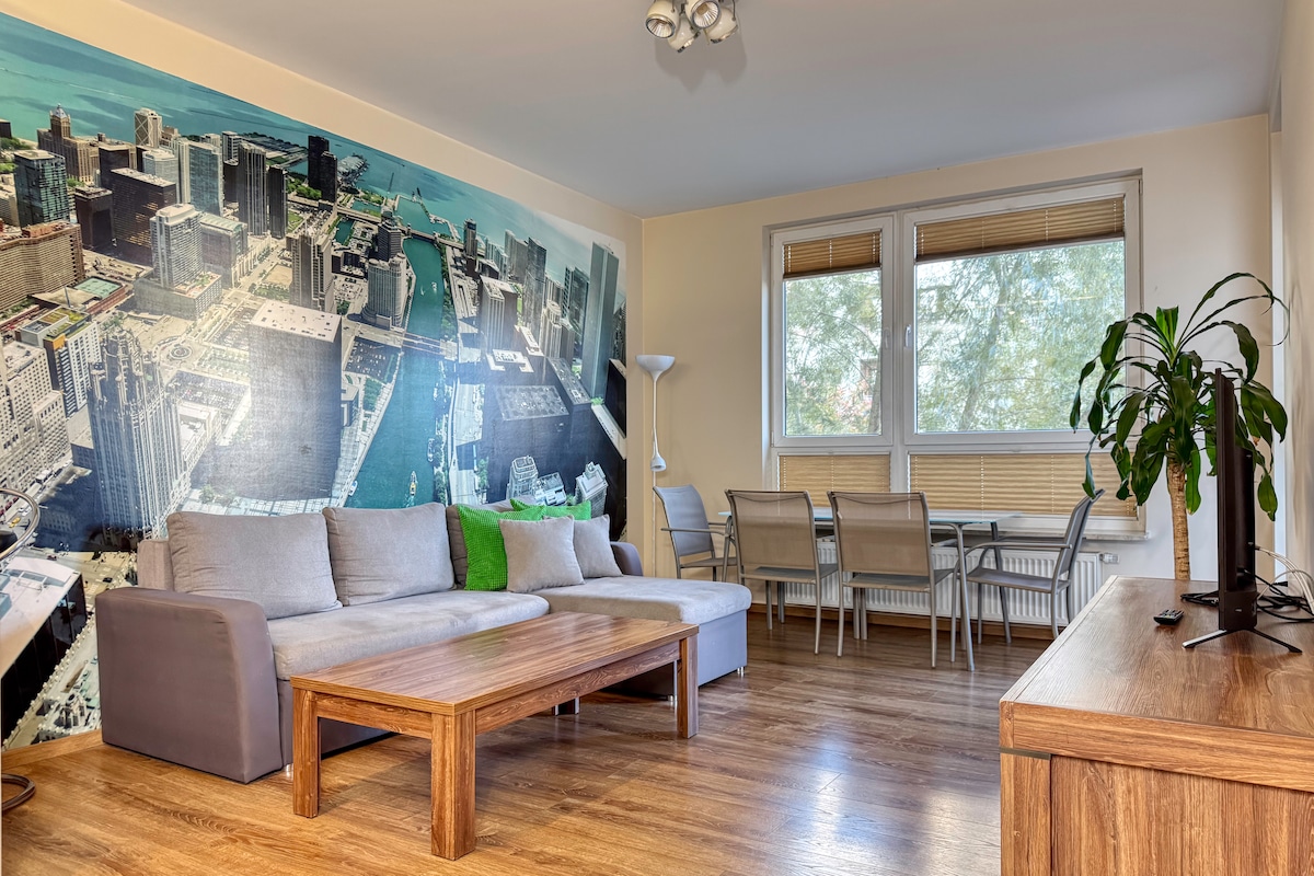 A spacious living area features a large couch and a wooden coffee table, complemented by a striking wall mural of a cityscape. Sunlight filters through large windows, illuminating a dining table with chairs nearby. A potted plant adds a touch of greenery to the inviting space.