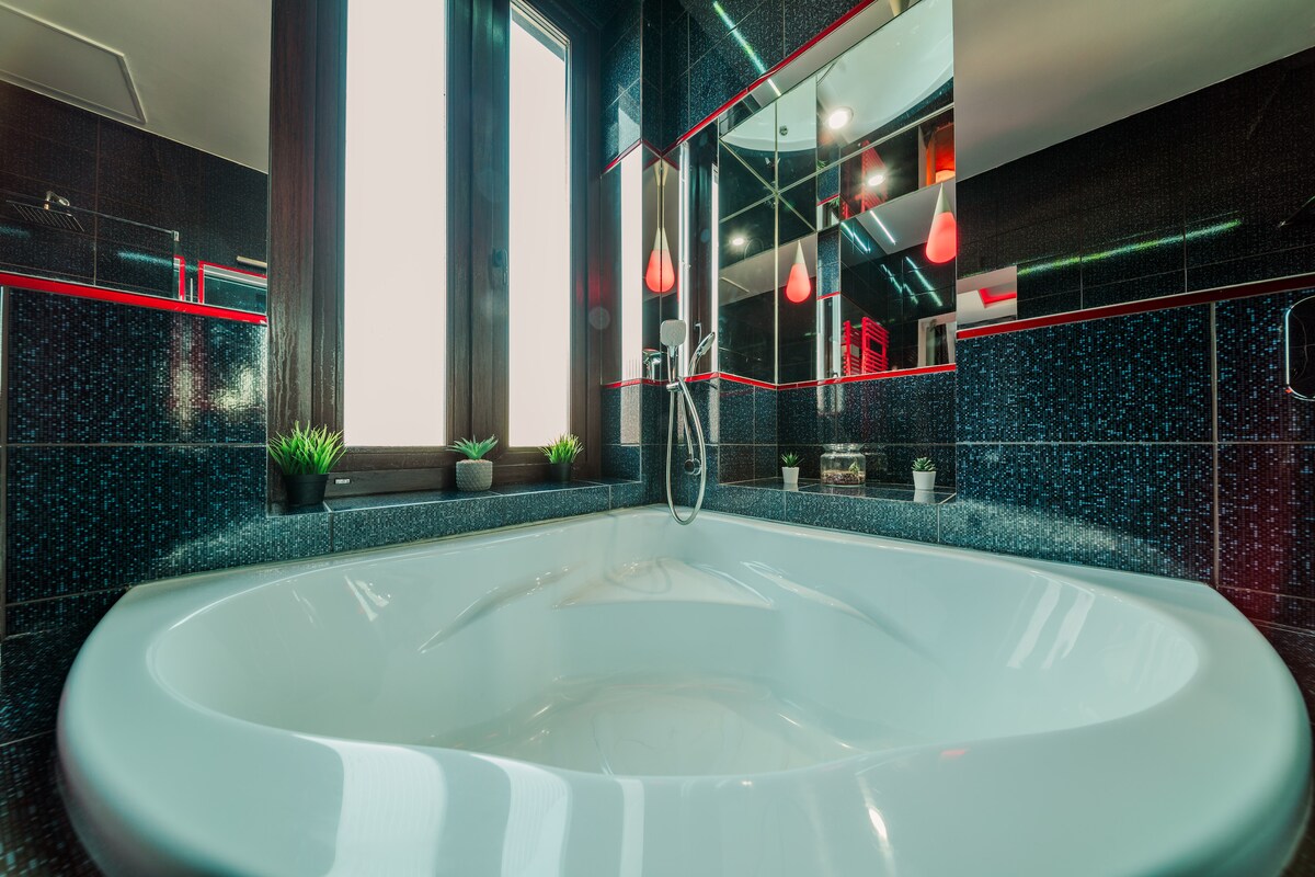 The bathroom features a spacious white hot tub, complemented by a wall of mirrors that enhance the light within the room. Decorative plants are placed on the counter, and ambient lighting highlights the sleek black tiles and red accents throughout.
