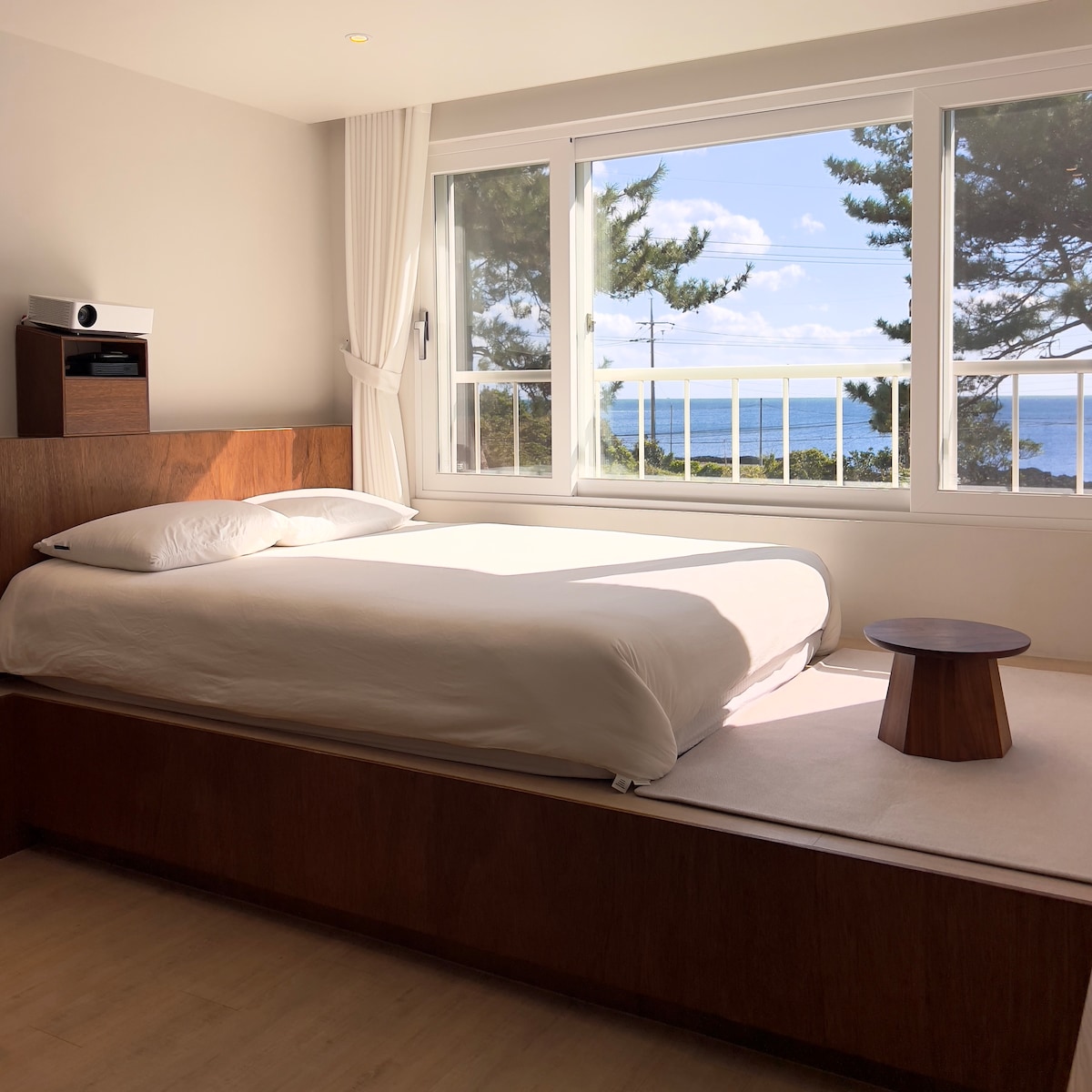 A spacious bedroom features a large bed with crisp white linens, positioned near a window offering a view of the ocean. Natural light fills the room, and a small round table is situated beside the bed. Soft colors create a serene ambiance.