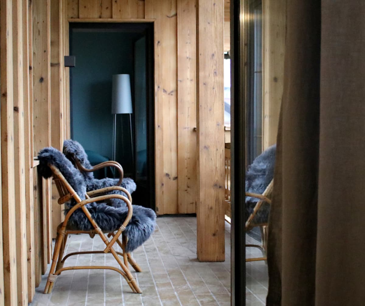 A hallway features wooden paneling with large windows, allowing natural light to fill the space. Two cozy chairs, adorned with soft gray throws, invite relaxation. A floor lamp stands in the corner, complementing the serene ambiance of the area.