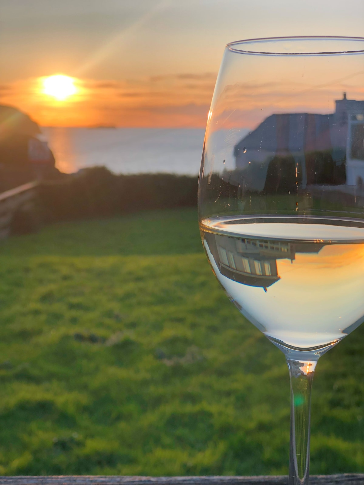 A glass of white wine is positioned in the foreground, reflecting the warm hues of a sunset over the sea. Soft grass and an outline of a building can be seen in the background, inviting a relaxed evening ambiance.