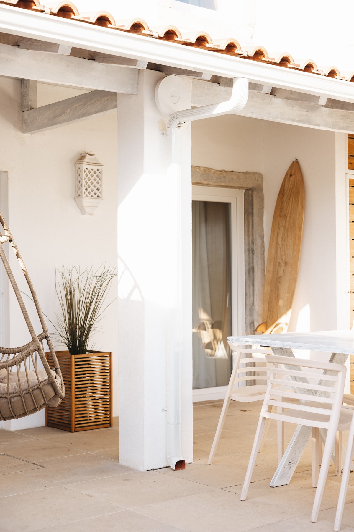 An airy terrace is featured, showcasing a minimalist dining table with white chairs. A cozy hanging chair is attached, complemented by potted plants and a wooden surfboard leaning against the wall. Sunlight brightens the space, highlighting the clean lines and natural materials.