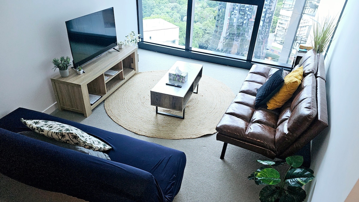 The living room features a comfortable brown sofa and a blue sofa bed, complemented by a wooden coffee table atop a round area rug. A large flat-screen television is positioned on a wooden console. Floor-to-ceiling windows provide a view of the surrounding greenery.
