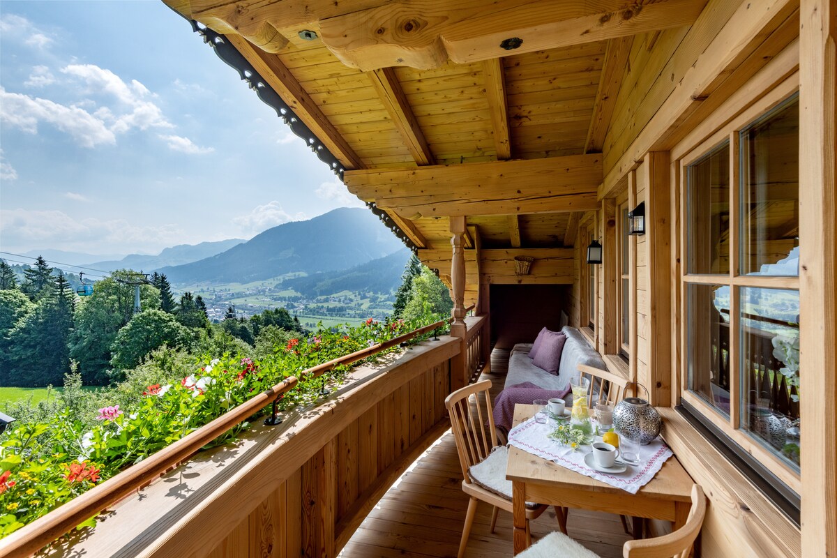 A wooden balcony offers a scenic view of the surrounding mountains and valley. A small table is set for breakfast, featuring a platter and beverages. Comfortable seating is arranged alongside vibrant flower planters, with sunlight illuminating the space and the warm wooden architecture.