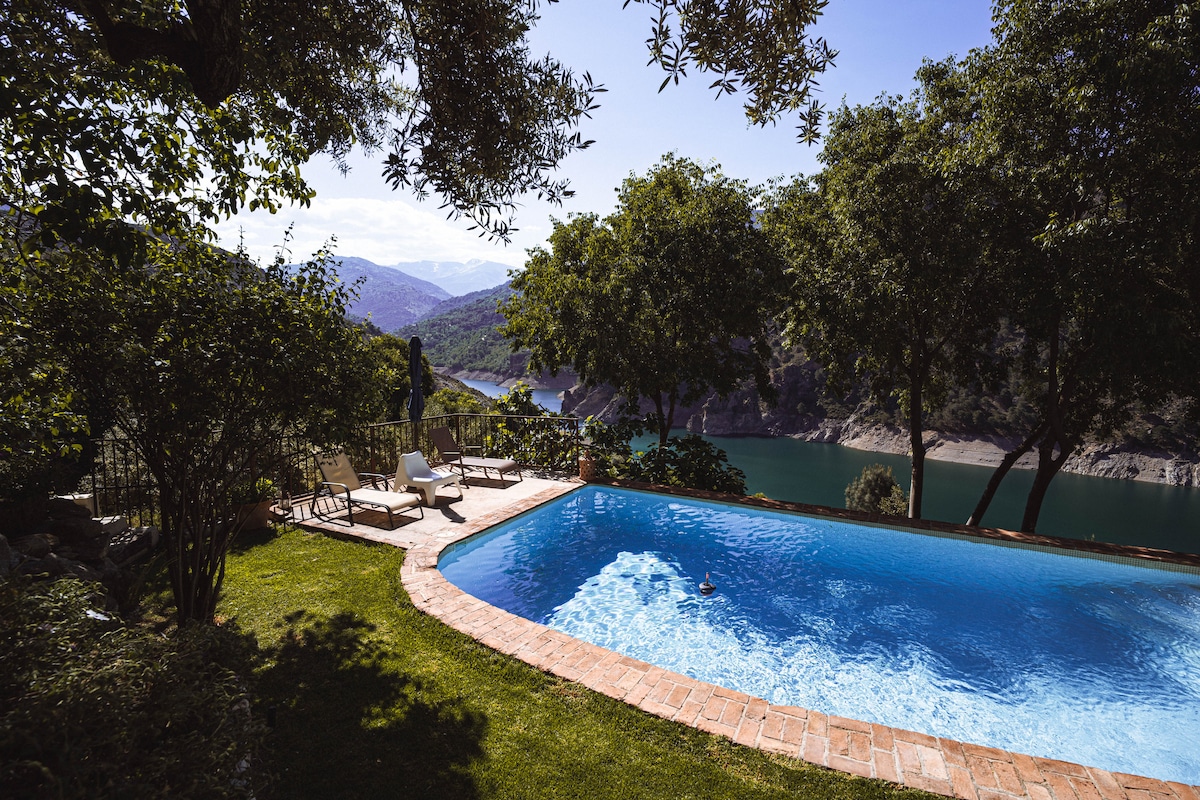 A private swimming pool is surrounded by lush greenery, offering a serene view of the Canales reservoir and the mountains beyond. Comfortable lounge chairs are placed near the pool for relaxation. The tranquil environment invites guests to enjoy the natural beauty of the area.