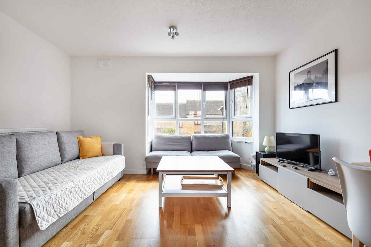 The living area features a comfortable grey sofa, complemented by a light grey sofa bed. A wooden coffee table sits centrally, with a television on a media unit nearby. Large windows allow natural light to fill the space, enhancing the wooden flooring.