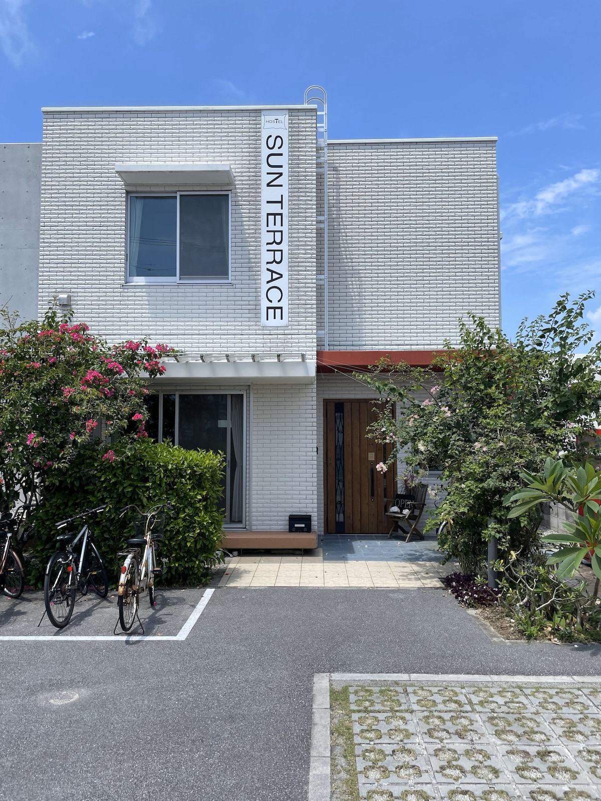 An exterior view of a modern, two-story building named 'SUN TERRACE' is shown. The structure features a bright white facade with a clear blue sky overhead. Lush greenery and pink flowering plants frame the entrance, enhancing the inviting appearance.