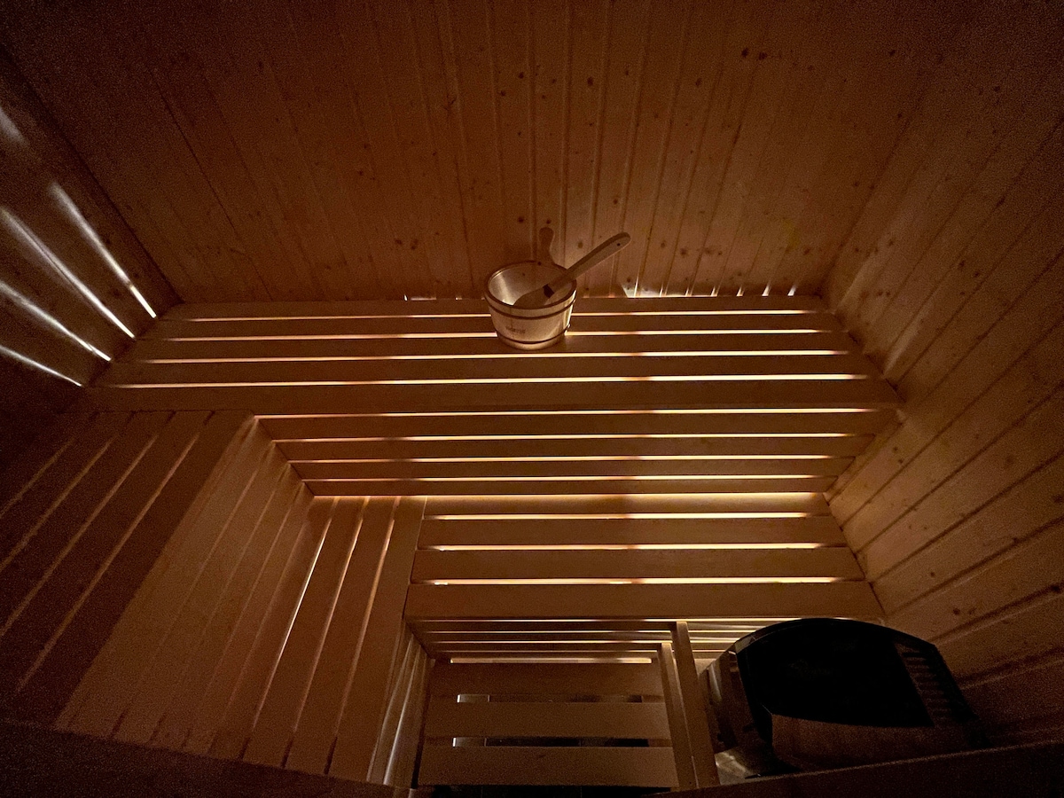 A sauna interior features warm wooden panels, with light filtering through slats, creating a serene atmosphere. A metal bucket is placed against the wall, and a wooden bench offers ample seating. An additional item is visible, enhancing the functional design of the space.