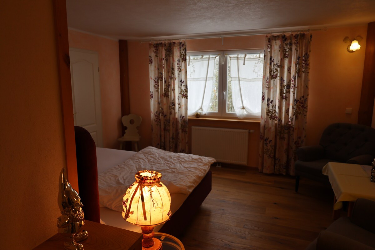 A warmly lit room features a spacious bed with light bedding and wooden flooring. Large windows adorned with floral curtains allow natural light to flow in. A cozy seating area includes a grey chair, while a decorative lamp adds a soft glow to the space.