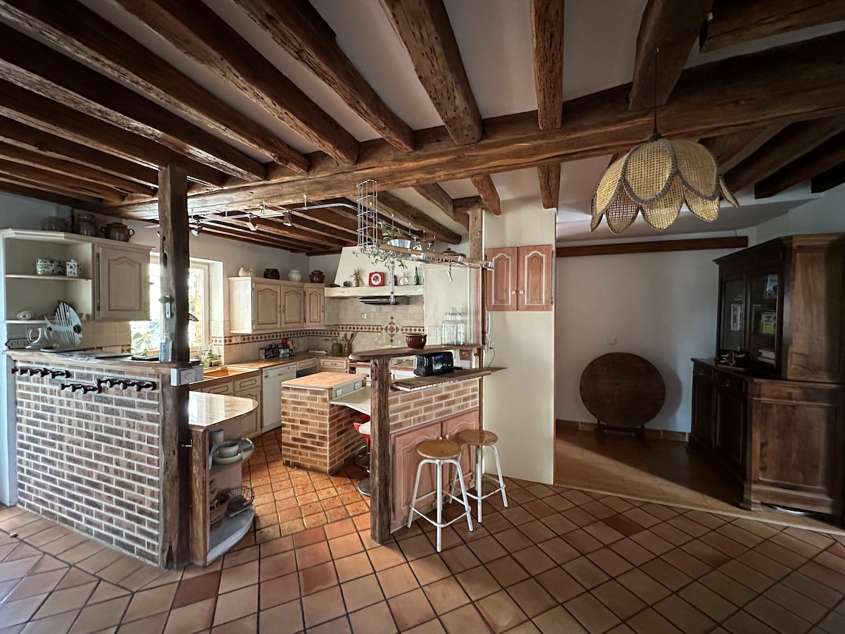 The kitchen features exposed wooden beams and a mix of traditional and modern design elements. A central island with barstools provides casual seating. Natural light enters through windows, highlighting brick accents and wooden cabinetry. A dining area is visible in the background.
