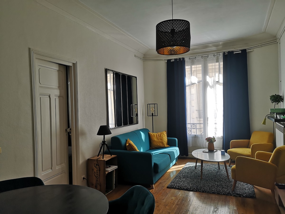 The living area features a comfortable blue sofa adorned with yellow cushions, complemented by a round coffee table on a textured rug. Two large windows allow natural light to illuminate the space, which includes a modern pendant light and decorative wall mirrors.