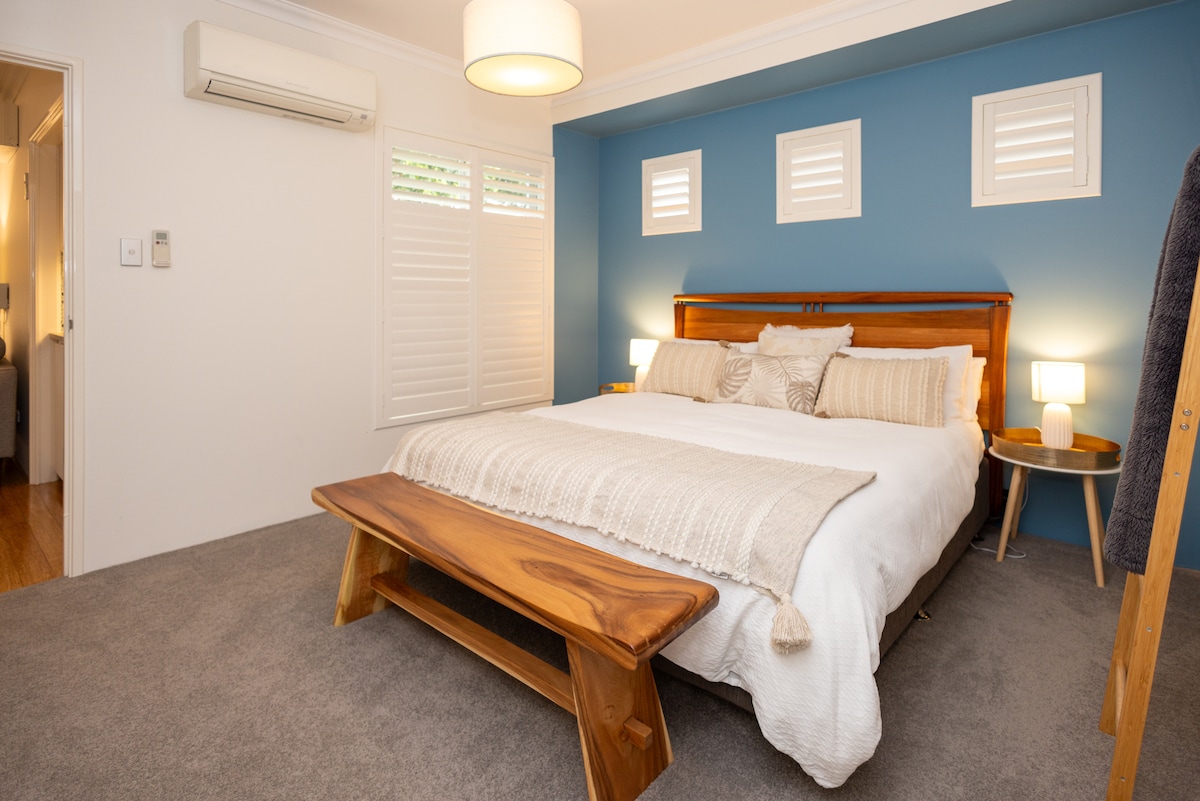 A cozy bedroom features a spacious bed adorned with neutral-toned linens. Natural light filters through small windows, highlighting the calming blue accent wall. A wooden bench sits at the foot of the bed, accompanied by a bedside table with a soft lamp. Air conditioning is discreetly positioned on the wall.