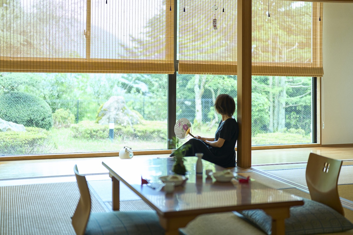 A serene living space features large windows draped with wooden blinds, offering a view of the garden. A person sits on the tatami floor near a low table, holding a fan. Natural light brightens the room, highlighting a tranquil setting.