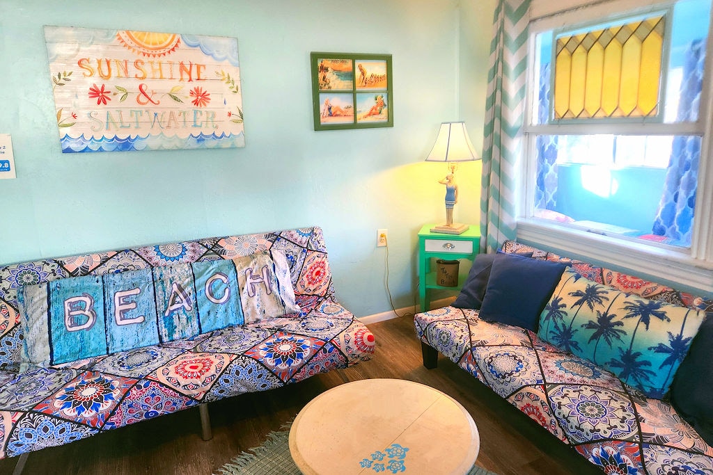 A welcoming living area features two patterned sofas adorned with cushions, emphasizing a relaxed beach theme. A round coffee table sits centrally, accompanied by a bright green side table with a lamp. Decorative wall art highlights the coastal atmosphere, with ample natural light streaming through a stained glass window.
