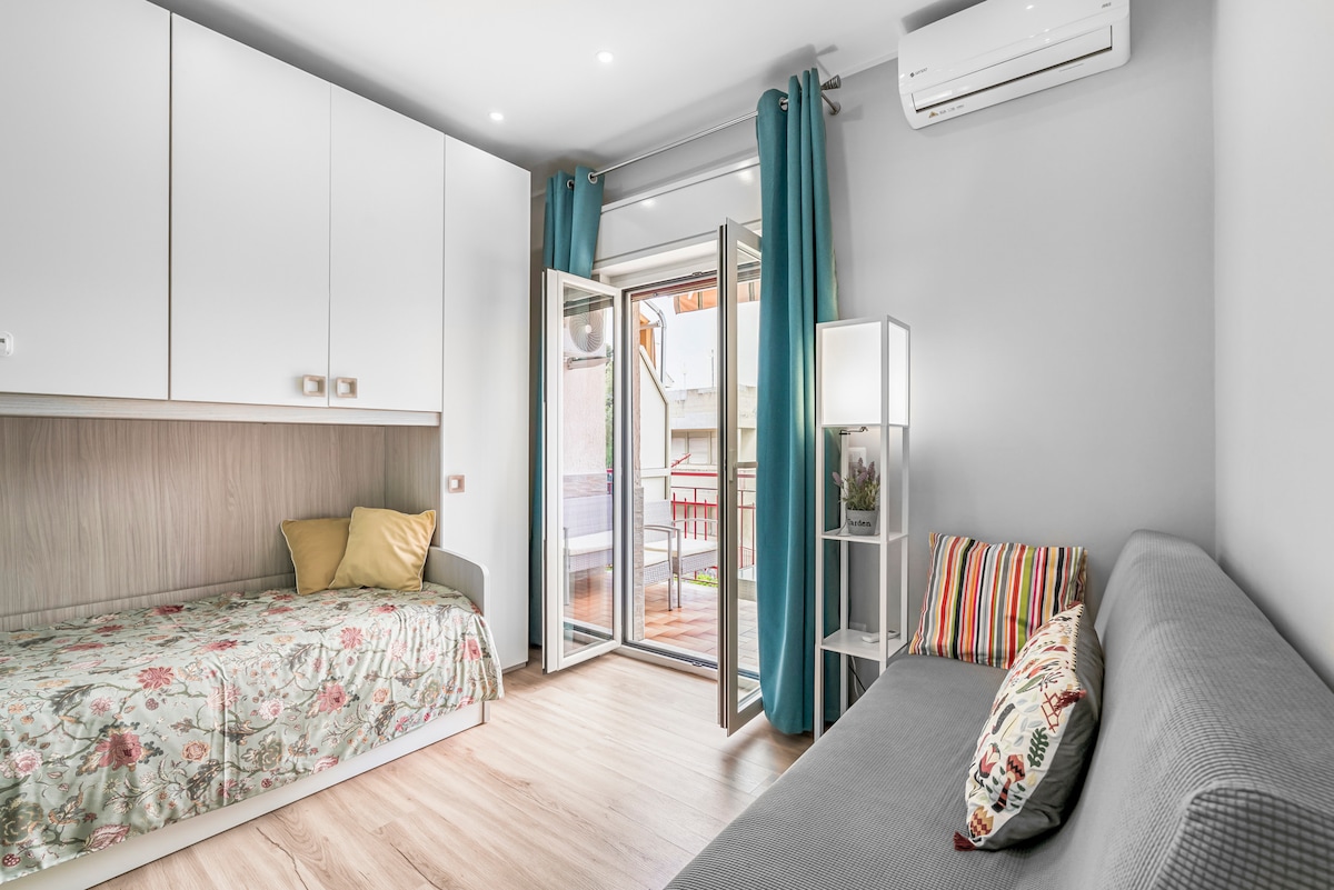 A cozy sitting area features a gray sofa, a single bed with a floral bedspread, and a decorative pillow. Large glass doors open to a private balcony, while light curtains and a stylish lamp add brightness to the space. Air conditioning is present for comfort.