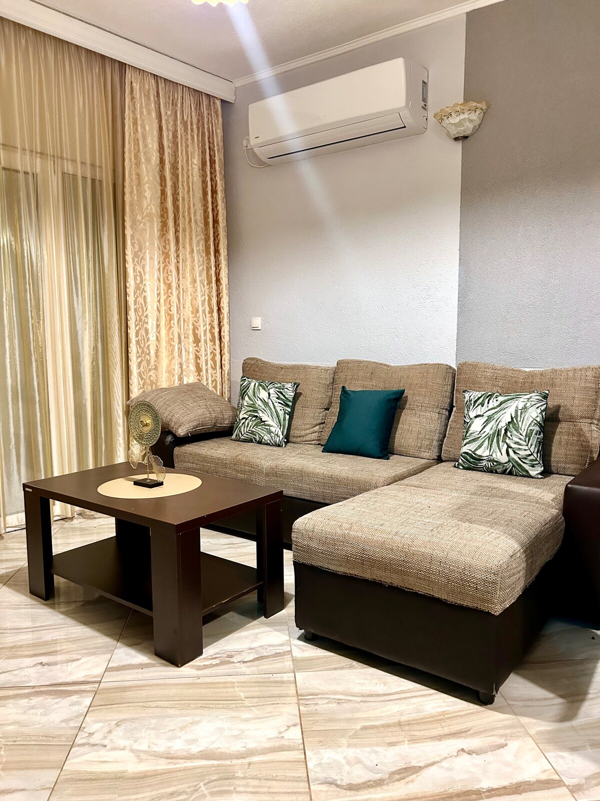 A comfortable living area is presented with a sectional sofa adorned with decorative cushions featuring tropical motifs. A small wooden coffee table sits in front of the sofa, and natural light filters through elegant sheer curtains, enhancing the space's inviting ambiance.