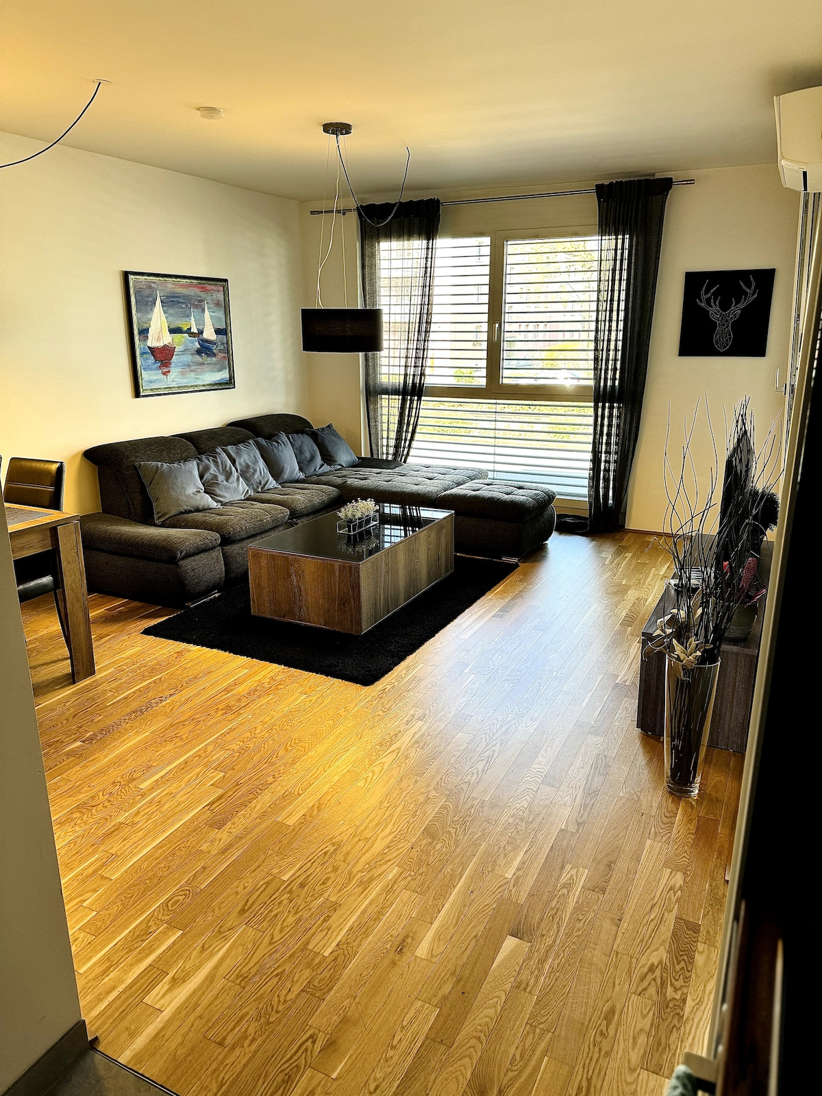 A living room features a comfortable dark sofa arranged around a wooden coffee table. Natural light filters through large windows adorned with sleek blinds. A vibrant wall art piece adds color to the space, while a dining table with chairs is visible in the background.