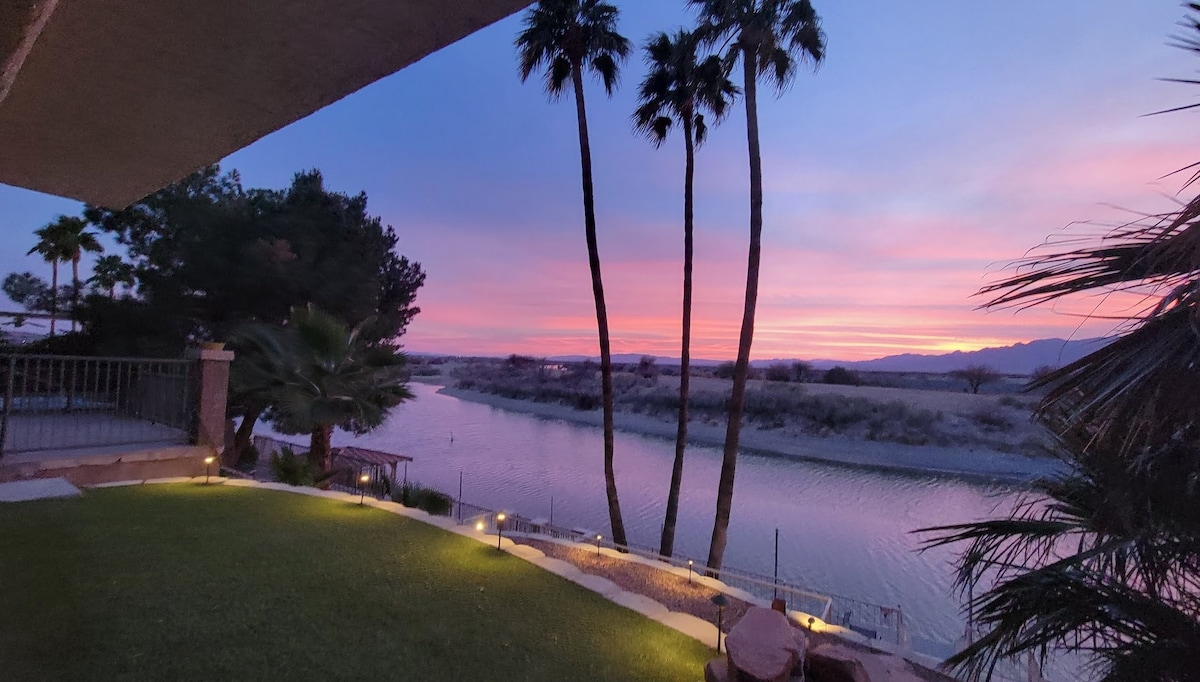 The image captures a scenic view of the Colorado River at sunset, showcasing a tranquil waterscape framed by palm trees. Soft hues of pink and purple illuminate the sky, while outdoor lighting accents the green grass and patio area, creating a peaceful atmosphere.