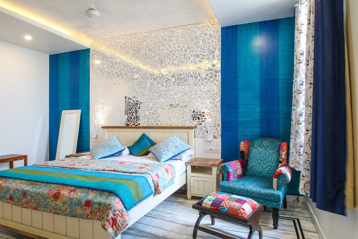 A spacious bedroom is characterized by vibrant blue and floral patterns, with a large bed adorned in colorful linens. A decorative wall feature adds texture to the space. A patterned armchair and a wooden table provide additional seating, complemented by natural light from the window.