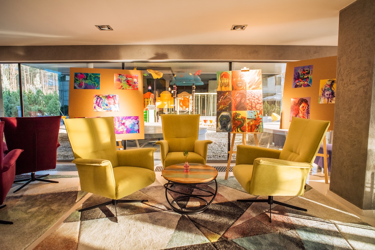 A cozy lounge area features four brightly colored chairs arranged around a round table. Art displays with colorful paintings are positioned in the background, while large windows allow natural light to fill the space. A geometric patterned rug adds texture to the floor.