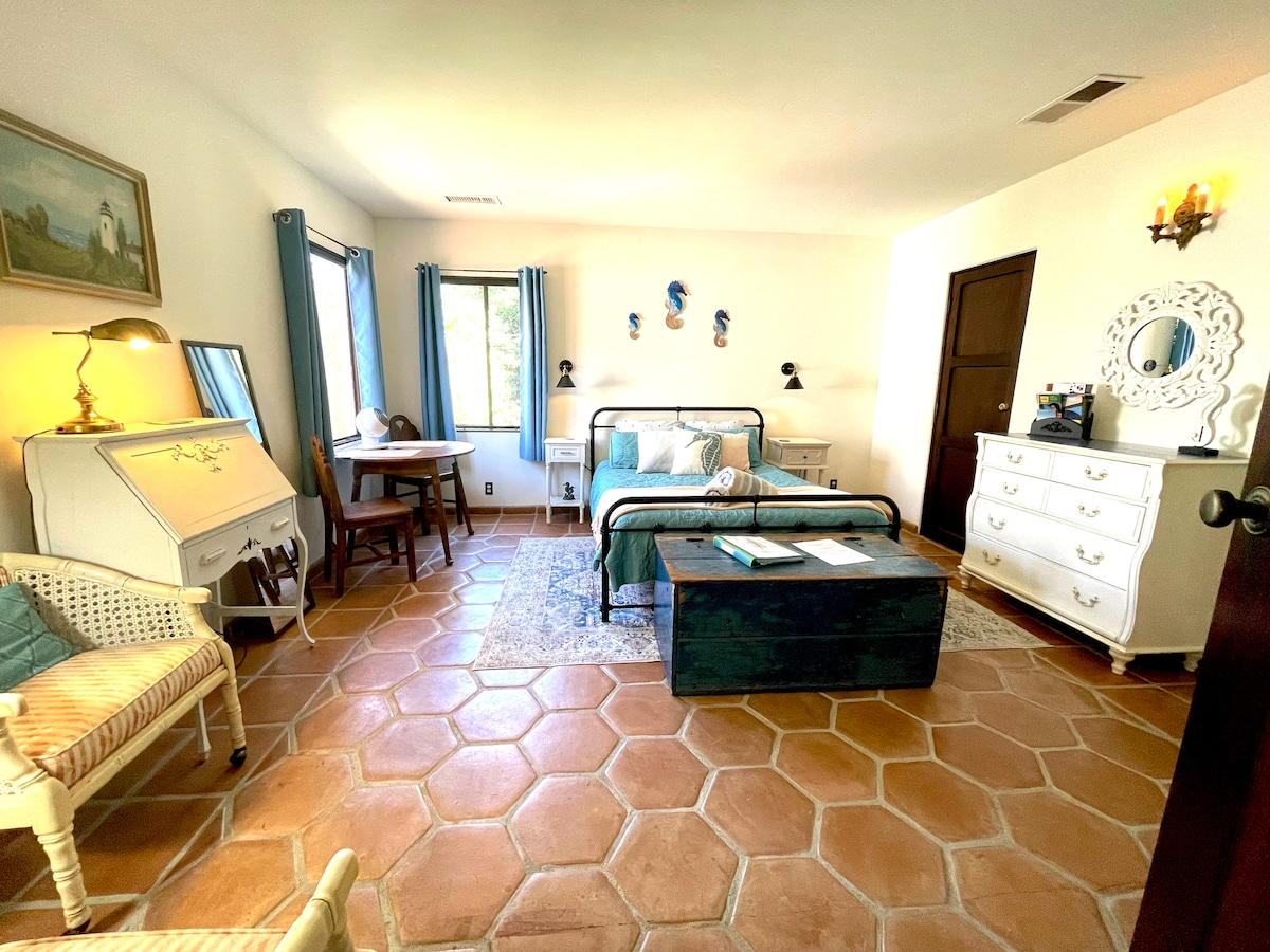A cozy guest room is highlighted by a spacious bed with a blue and white color scheme. Soft lighting emanates from wall sconces. A small table and chairs offer a dining area, while rustic tiled flooring adds warmth. A vintage dresser complements the decor.