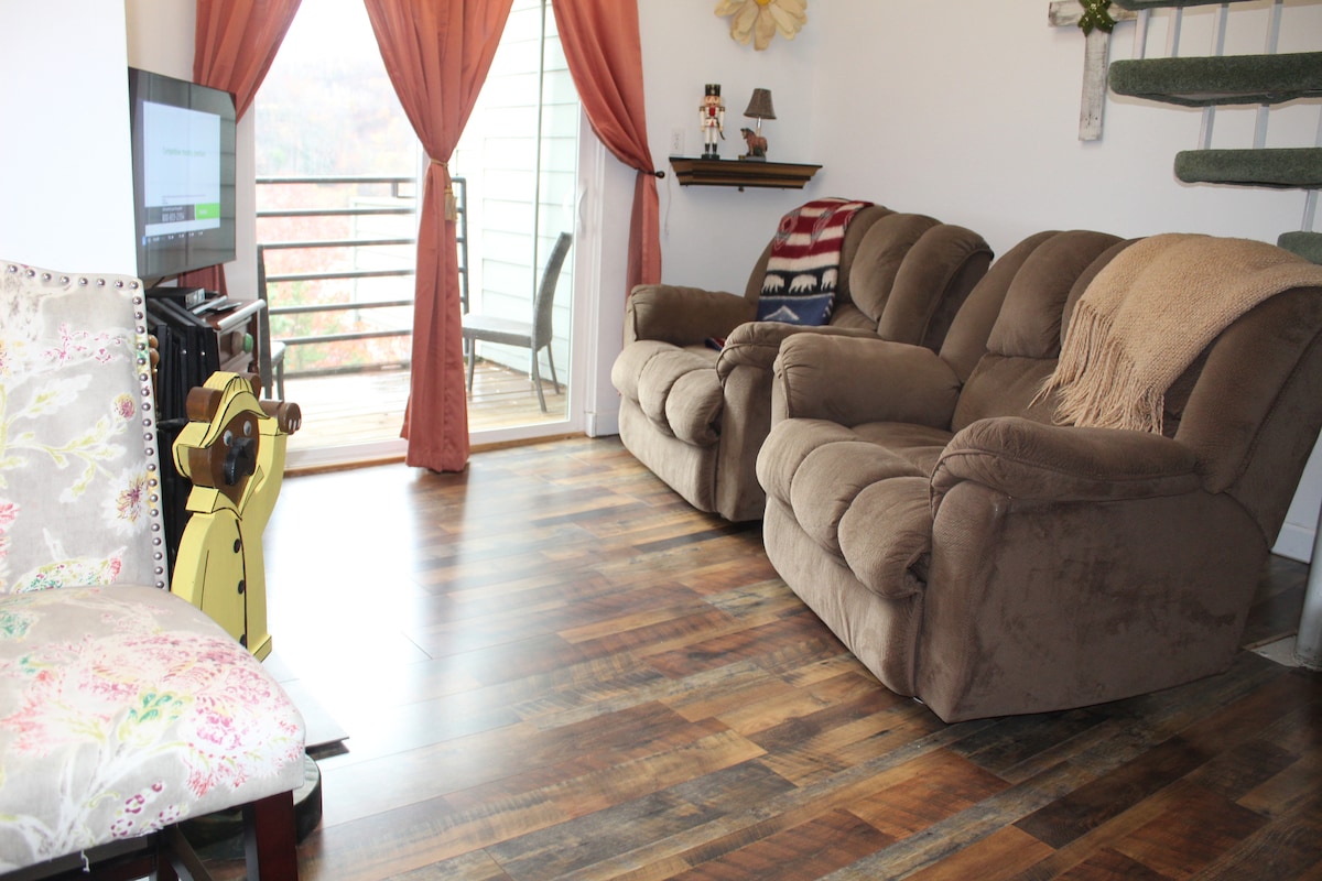 The living area is furnished with two comfortable recliners and a floral accent chair. A flat-screen TV is positioned on a stand, and large windows, framed by curtain panels, offer views of the balcony and surrounding nature.