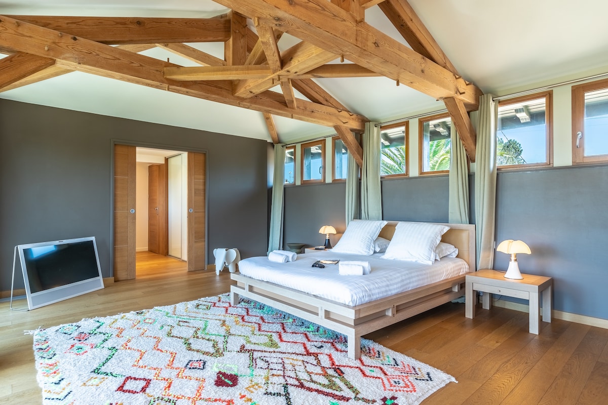 A spacious bedroom features a large bed with fresh linen, complemented by bedside tables and modern lamps. Expansive windows allow natural light to fill the room, highlighting the wooden beams that provide architectural interest. A colorful area rug adds warmth to the wooden floor.