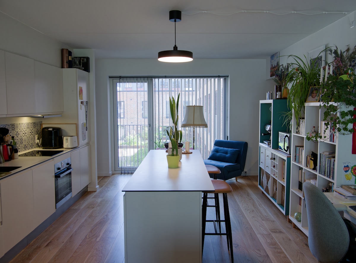 The kitchen area is bright and airy, featuring a long dining table with bar stools. A bay window allows natural light to flood in, showcasing potted plants and a cozy armchair. Shelves hold decorative items, enhancing the modern atmosphere.
