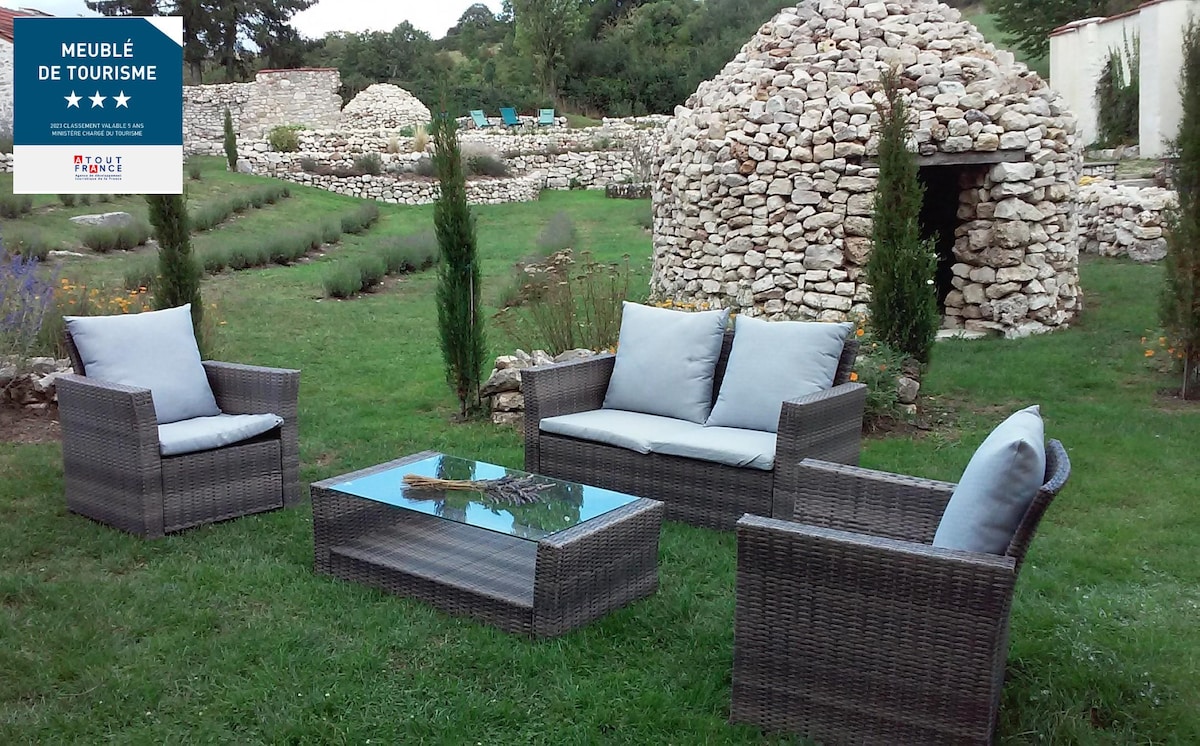 An outdoor seating area is presented with a modern design, featuring a glass table surrounded by four comfortable wicker chairs with light-colored cushions. In the background, stacked stone structures and a landscaped garden with greenery and lavender complete the tranquil setting.
