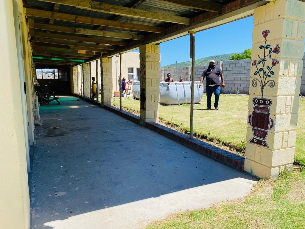 A covered outdoor area features a concrete floor and brick columns, offering a view of the grassy yard. A decorative mural of flowers is visible on one wall. People are seen in the distance, while a round structure is placed in the yard.
