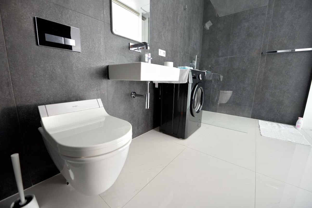 A modern bathroom features sleek dark walls and a bright floor. A wall-mounted sink and a toilet are positioned adjacent to each other, with a washing machine included. A large mirror reflects the well-lit space, enhancing the clean and contemporary design.