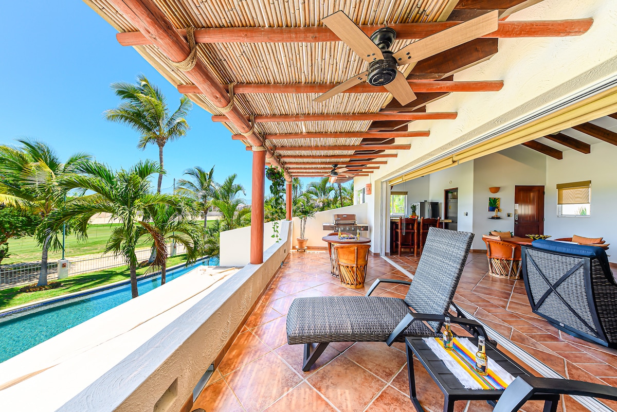 A spacious terrace is featured with comfortable lounge chairs and a ceiling fan, under a thatched roof. Palm trees gently sway in the breeze, while a view of the pool and golf course is visible in the background, contributing to a relaxed atmosphere.