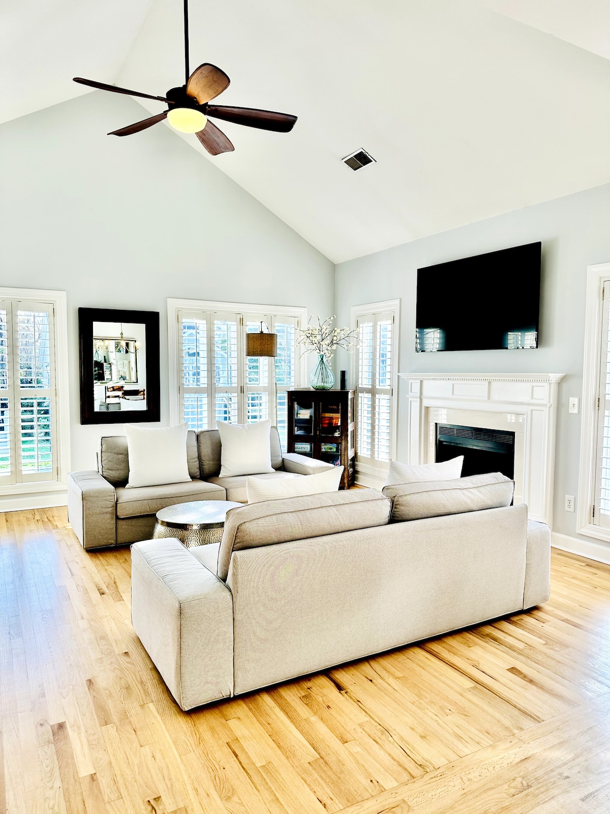 A bright living room features a sectional sofa arranged around a glass-topped table. A ceiling fan hovers above, with natural light streaming through several shuttered windows. A fireplace is situated beneath a mounted television, enhancing the room's inviting and functional layout.