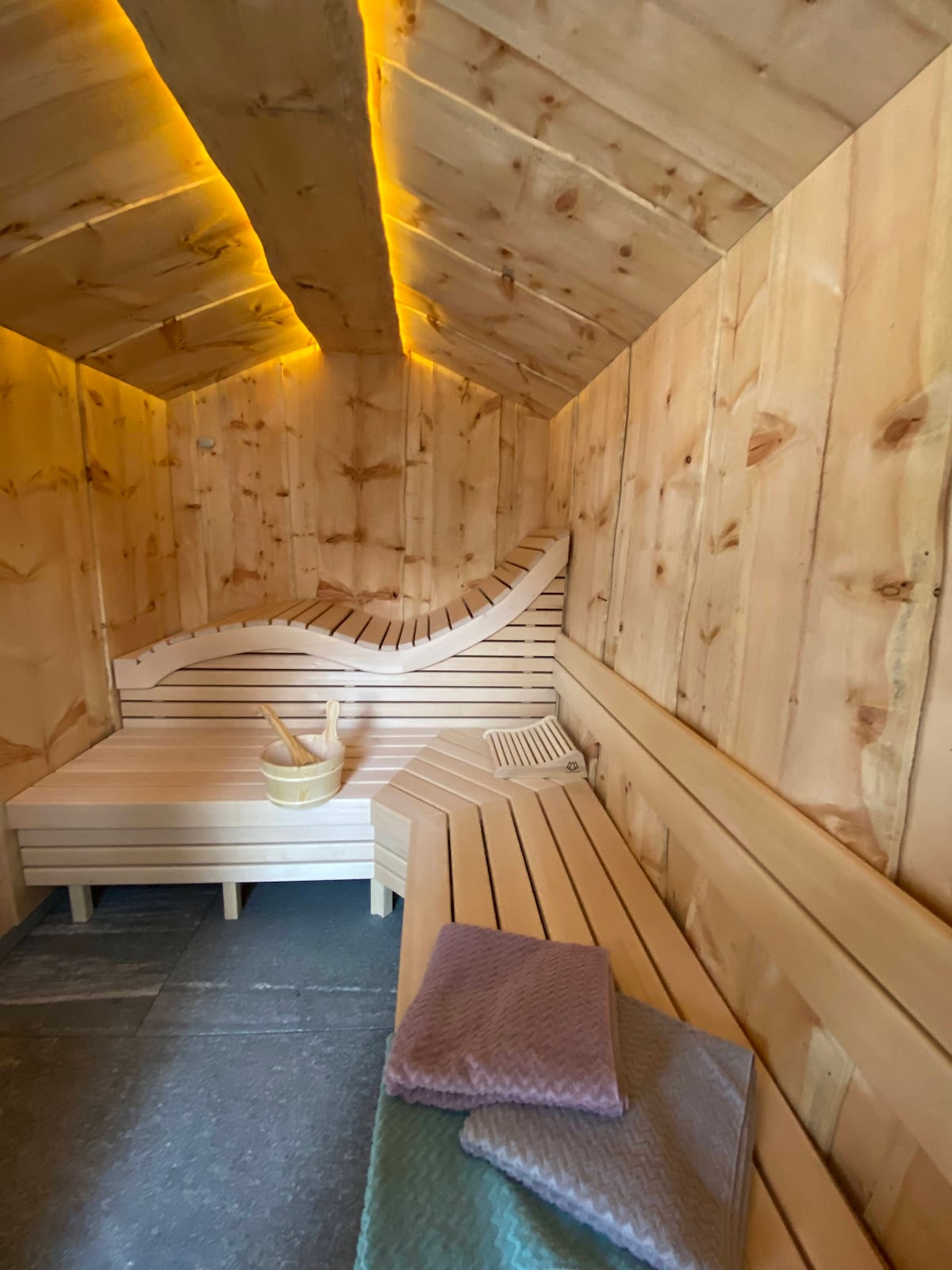 A warm and inviting sauna is surrounded by smooth wooden walls and ceiling, creating a serene atmosphere. It features curved wooden benches with a slatted design, a small wooden bucket, and a towel resting on one of the benches.