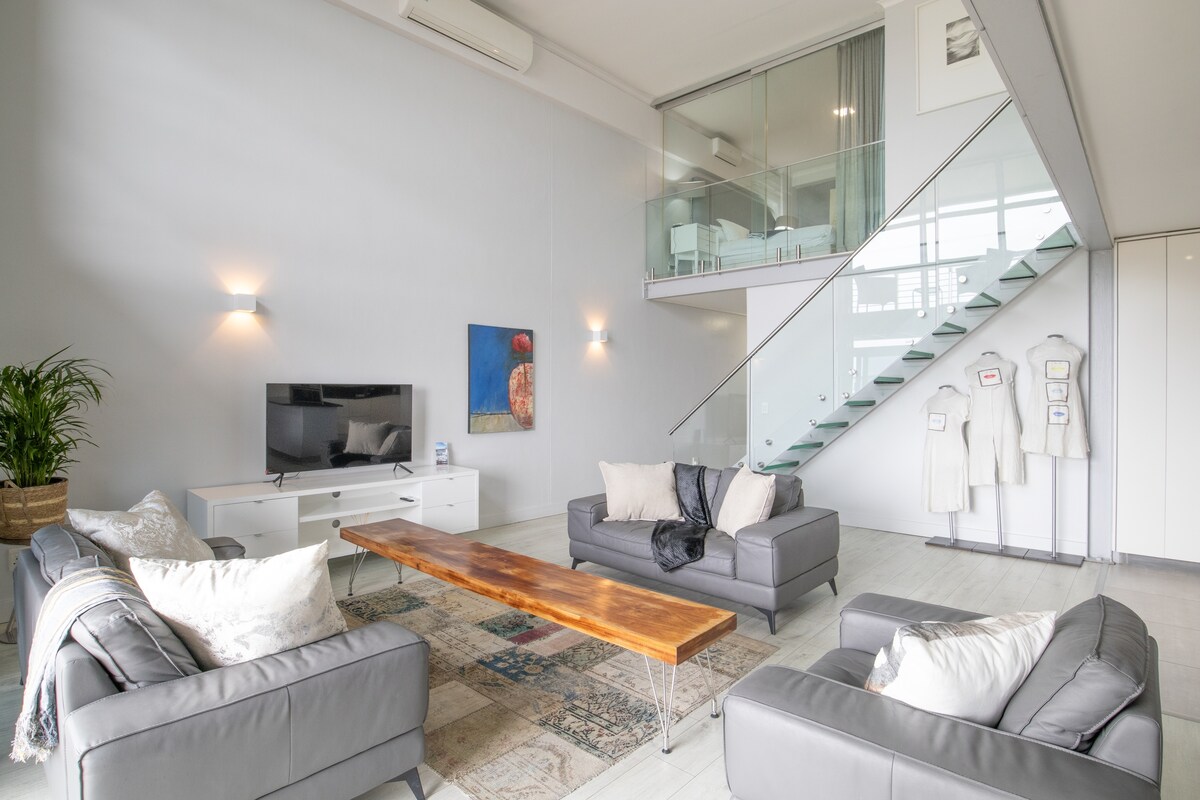 An open-plan lounge area features modern leather couches oriented around a long wooden coffee table. Natural light pours in through large windows, while a minimalist television unit sits against the wall. A staircase with glass railings leads to the lofted area above.