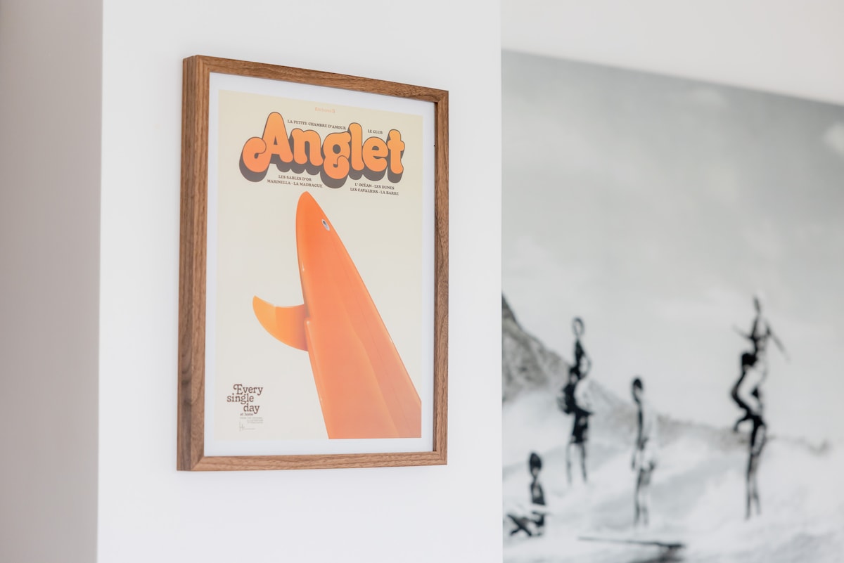 A framed poster displaying the word 'Anglet' is mounted on a white wall. The orange surfboard featured in the artwork adds a vibrant touch. A black-and-white photograph of people surfing is visible in the background, contributing to the coastal theme.