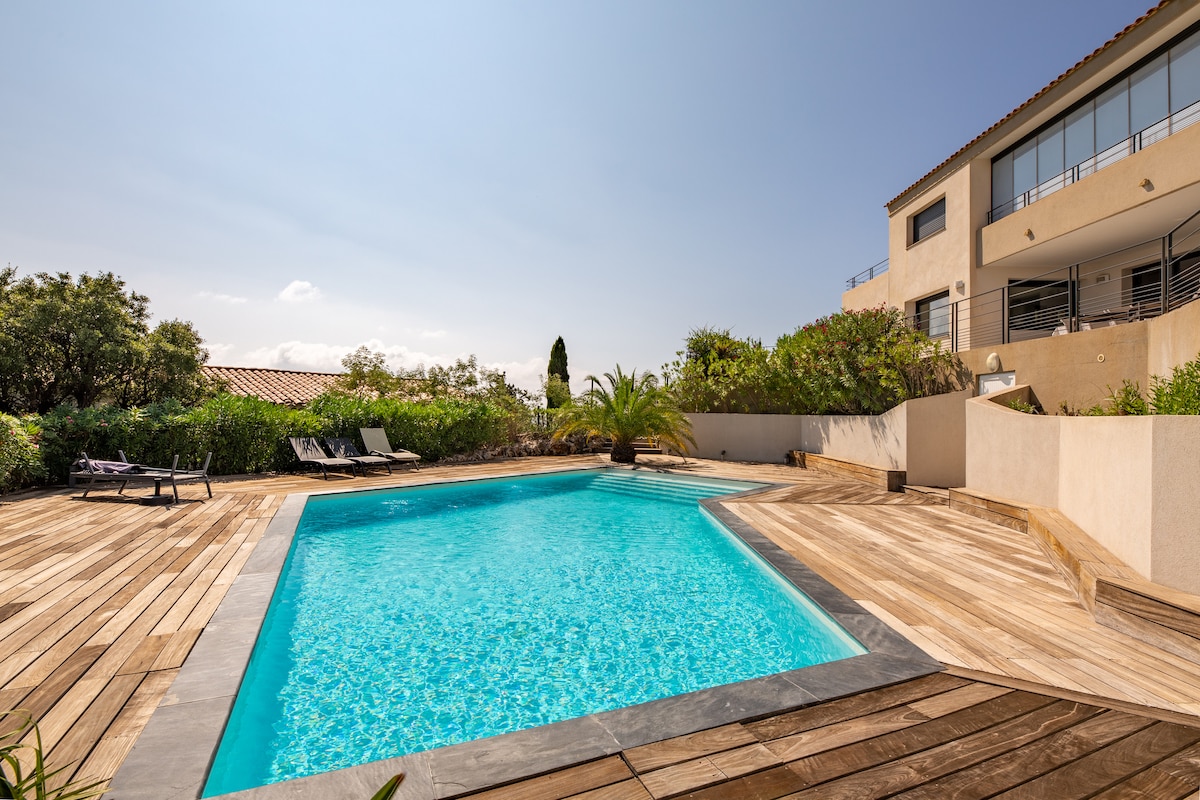 The image displays a modern pool surrounded by a wooden deck, featuring lounge chairs placed for sunbathing. Lush greenery and shrubbery create a private atmosphere. The building's contemporary architecture is visible in the background, with clear skies overhead contributing to the serene environment.