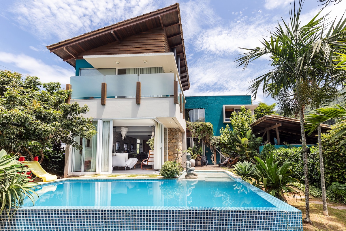 A modern house with a spacious outdoor area is presented, featuring a private pool with a vibrant blue hue. Lush greenery surrounds the property, and large glass doors create a seamless connection between indoor and outdoor spaces. The upper level showcases a wooden roof and inviting balcony.