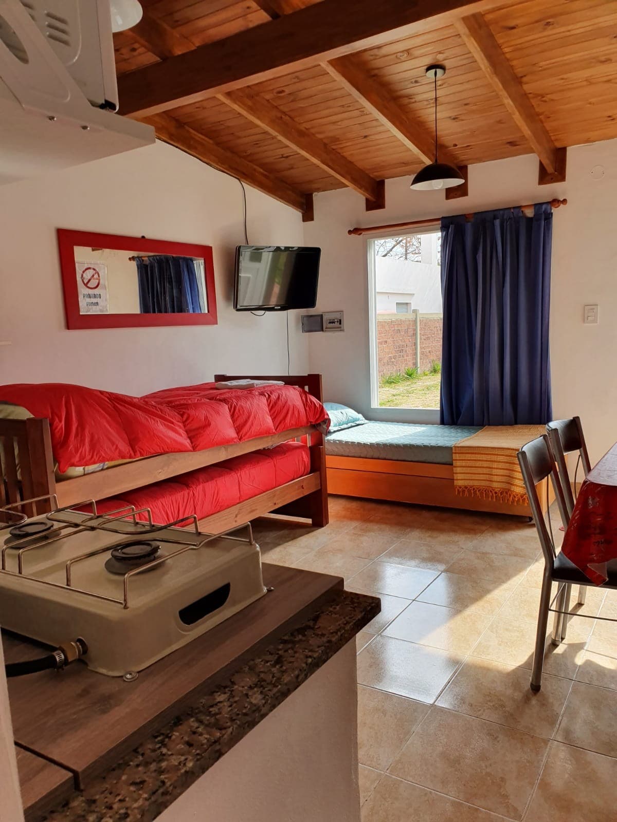 A bright room is seen featuring a spacious bed with a red comforter and a separate mattress. A window with dark curtains allows natural light to enter, and a television is mounted on the wall. A dining table and stove are positioned in the corner, contributing to the functional layout.