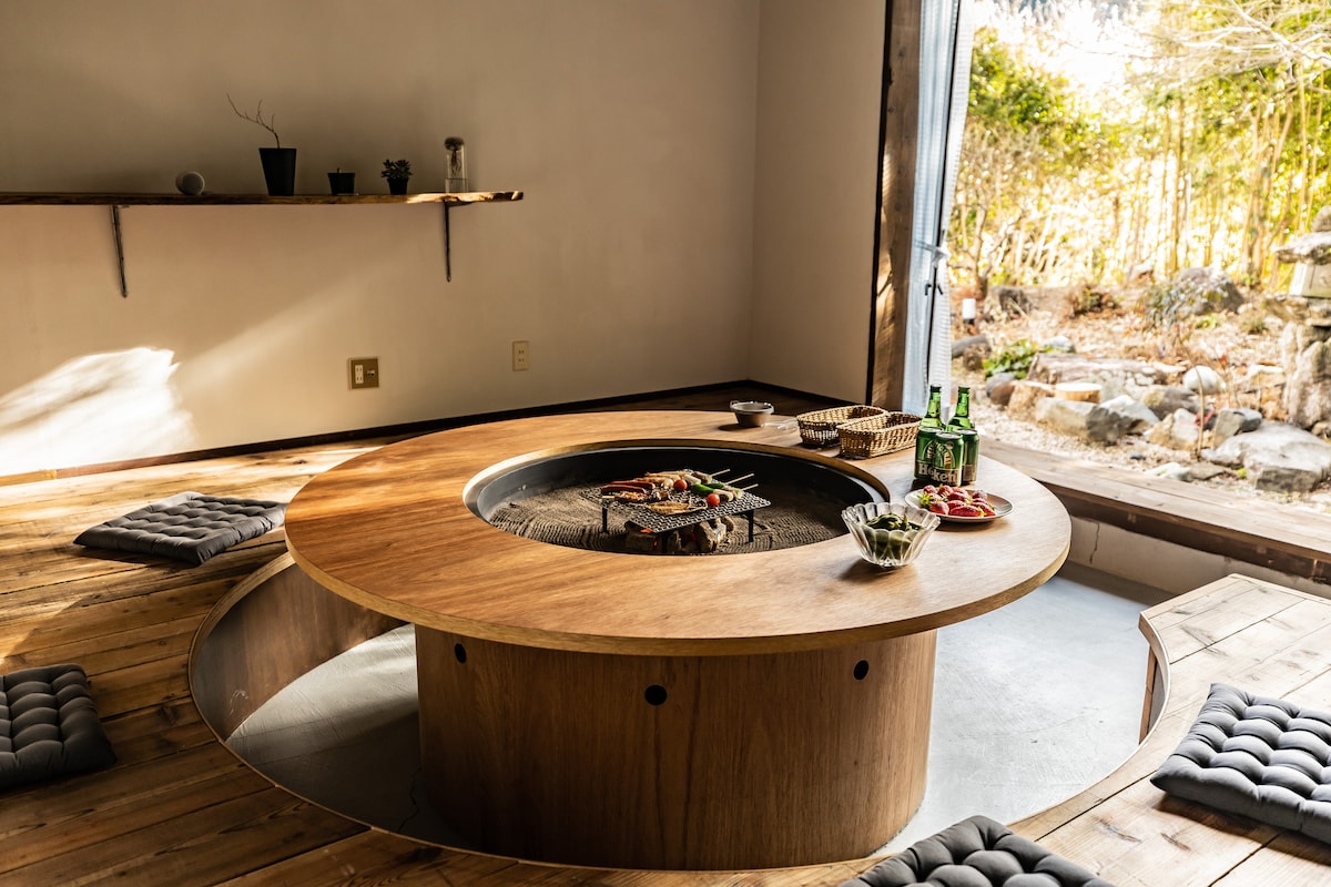 An inviting indoor space features a round wooden table with a built-in irori (traditional Japanese hearth) at its center. Comfortable seating cushions surround the table, while soft natural light filters through an open doorway, showcasing a serene outdoor garden view.