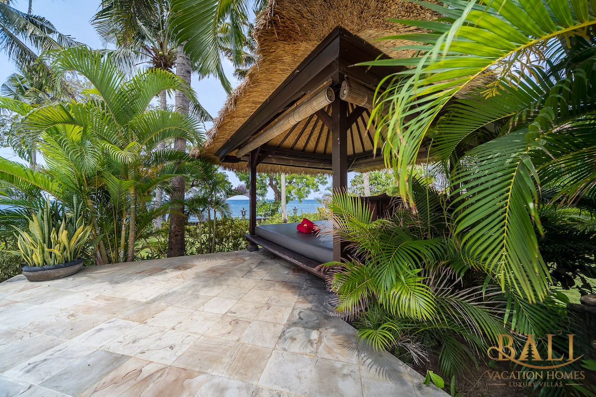 A Balinese relaxation bale is nestled among tropical plants, providing a shaded area for lounging. The spacious platform features a cushioned mattress, inviting guests to unwind while enjoying views of the ocean and surrounding greenery.