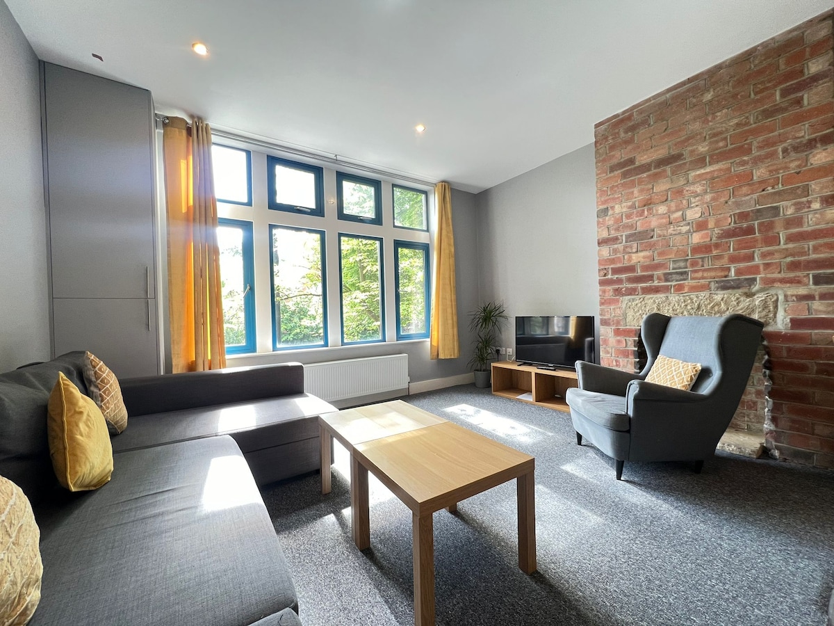 The living area is filled with natural light from large windows adorned with yellow curtains. A comfortable sofa faces a wooden coffee table, with a simple armchair positioned nearby. A flat-screen TV rests on a low cabinet against a feature brick wall.