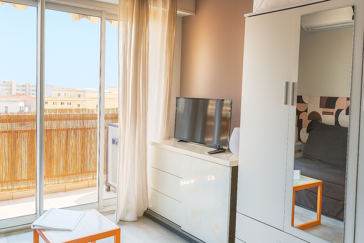 A light-filled studio features a minimalist design with a large window and sliding door leading to a balcony. A television sits atop a white dresser, while a cozy corner with a spacious bed is visible. Sheer curtains frame the window, enhancing natural light.