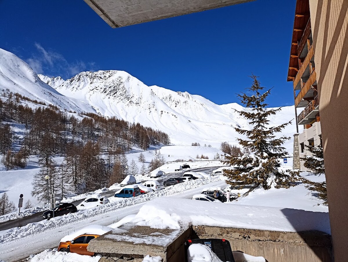 A view of snow-covered mountains under a clear blue sky is captured from the apartment's balcony. Vehicles are parked along a snowy road, with trees scattered across the landscape, enhancing the serene winter atmosphere.