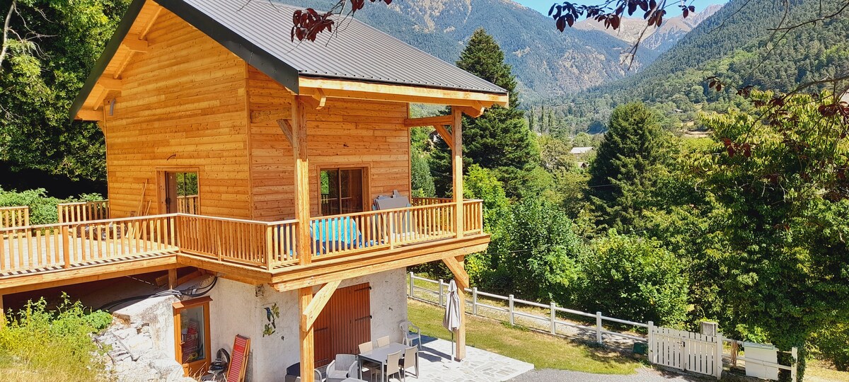The exterior of a wooden chalet is presented, featuring a spacious balcony with seating. Surrounding greenery and mountains create a serene backdrop. A patio area with chairs is visible, inviting outdoor relaxation. The structure demonstrates a blend of natural materials and contemporary design.