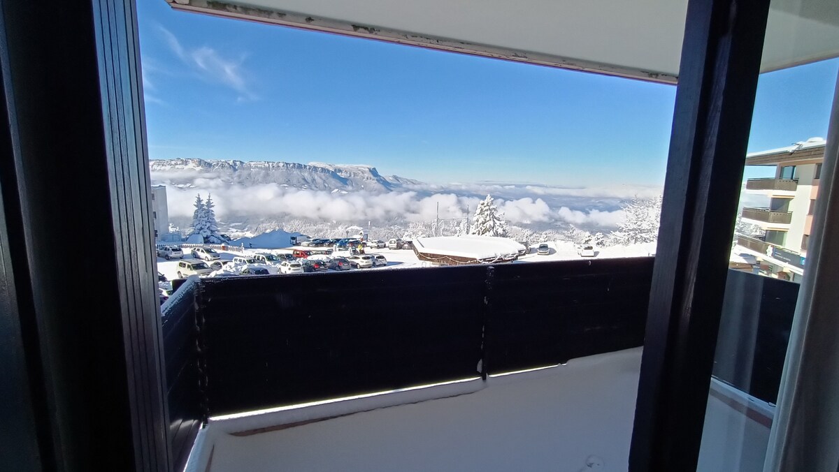 A balcony view showcases a winter landscape, featuring snow-capped mountains and clouds below. Vehicles are parked in the foreground, with a natural scene of trees and peaks in the distance, creating a serene atmosphere. The clear blue sky adds to the tranquil setting.