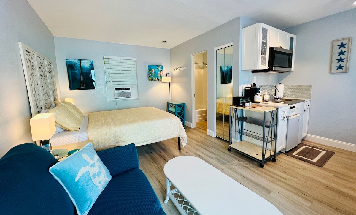 A light-filled studio features a spacious queen bed with a soft yellow duvet, complemented by coastal-themed decor. A kitchenette with modern appliances and a dining table is visible, alongside a comfortable blue sofa and a coffee table, creating a welcoming living space.