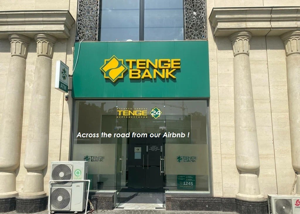 The exterior of Tenge Bank is displayed, featuring a prominent green sign with the bank's logo. Large glass doors offer a view into the bank's interior. Air conditioning units are positioned beside the entrance, with decorative stone columns flanking the building.