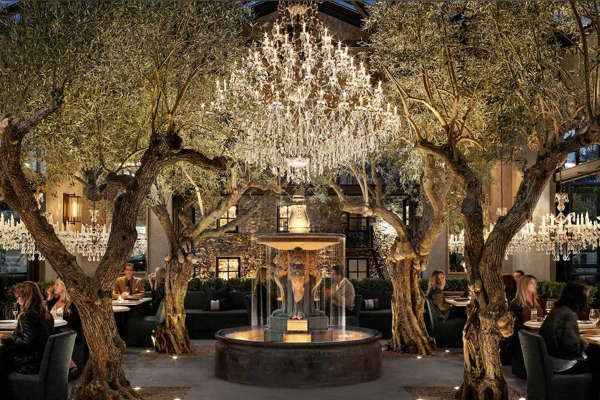 An elegant dining area is surrounded by olive trees and adorned with sparkling chandeliers. A central fountain reflects ambient light, creating a tranquil atmosphere. Guests are seated at tables, enjoying their meals beneath the canopy of shimmering lights.
