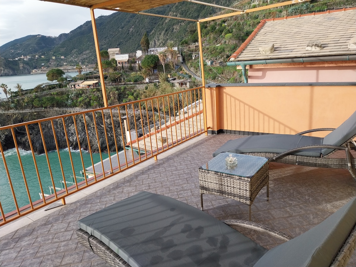 A terrace area features lounge chairs and a small glass table, offering views of the coastline and surrounding hills. The railing provides safety while allowing an uninterrupted sight of the sea and landscape, creating a quiet outdoor space for relaxation.
